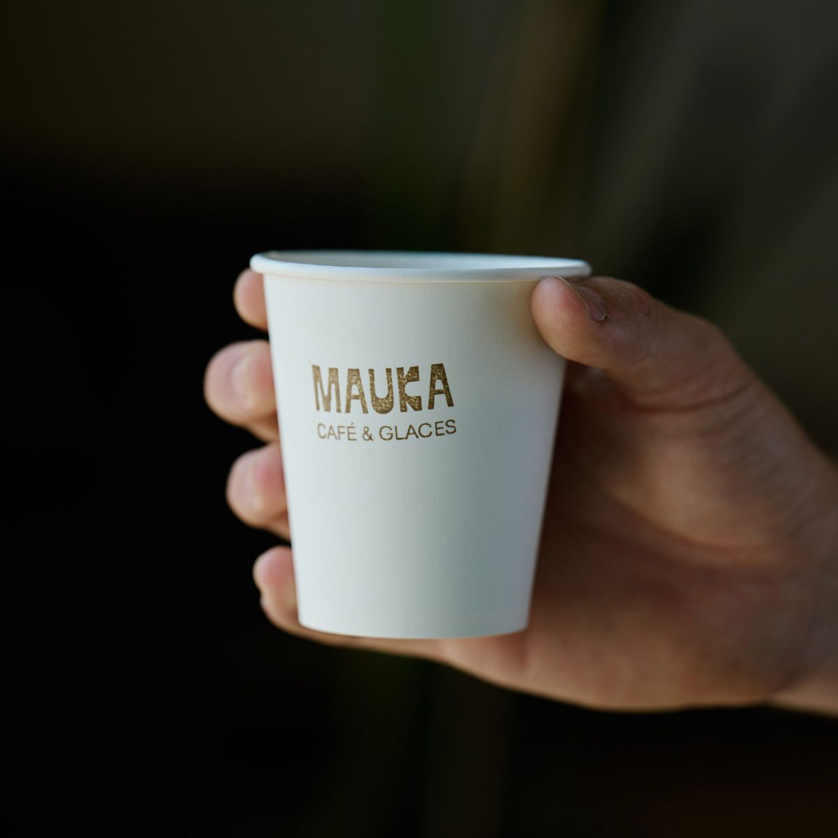 Hand holding a Mauka Café & Glaces paper cup with gold logotype